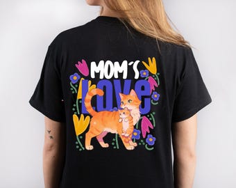 Puede incluir: Camiseta negra con una ilustración colorida de una gata y su gatito con las palabras "MOM'S LOVE" en blanco y azul. El diseño incluye flores en rosa, amarillo y azul.