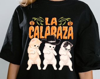 Può includere: Una t-shirt nera con il testo arancione LA CALABAZA e due piccole icone di zucca. Sotto il testo ci sono tre gattini: uno arancione chiaro, uno bianco con un cappello da cowboy nero e uno bianco e nero con occhiali rotondi.