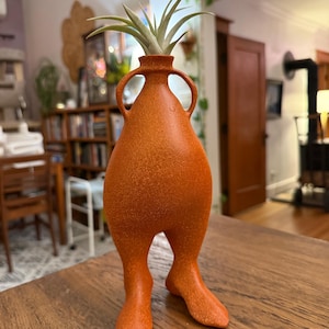 May include: Orange ceramic vase with a textured surface, featuring a unique anthropomorphic design with legs and handles. The vase holds a green air plant. The vase is on a wooden table.