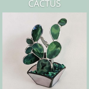 May include: A three-dimensional glass cactus sculpture with green glass leaves and a silver frame, set in a pink and white pot filled with green stones. The text "3 DIMENSIONAL CACTUS" and "SAGE GLASSWORK" are displayed.