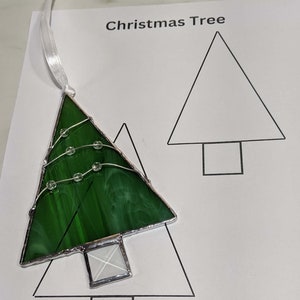 Puede incluir: Adorno de árbol de Navidad de vidrio verde con contorno de alambre plateado y detalles de cuentas transparentes. Tiene una cinta blanca para colgar. Las palabras "Christmas Tree" y "SageGlasswork.com" están impresas en el papel.