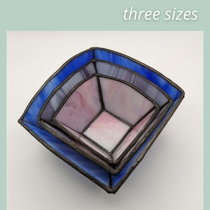 May include: Three square, stained glass stacking bowls in shades of blue, lavender, and pink. The bowls are nested, with the largest bowl in a vibrant blue. The text "STACKING BOWLS three sizes" is at the top.