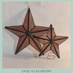 May include: Two stained glass 3D five-pointed stars in shades of mauve and brown, with a dark border. The larger star has a loop for hanging. Text reads "3D FIVE POINTED STAR" and "2 sizes". The bottom text says "SAGE GLASSWORK".