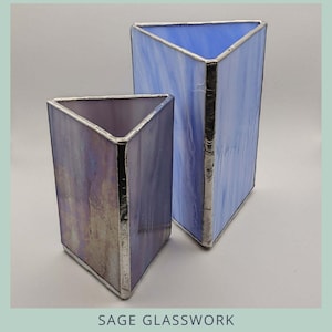 Puede incluir: Dos jarrones triangulares de cristal de diferentes tamaños. El jarrón más grande es azul claro y el más pequeño es morado. Ambos tienen ribetes plateados y la inscripción "Triangle Vase" y "two sizes".