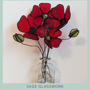 May include: A stained glass poppy seedhead and flower arrangement in a clear glass vase. The red flowers have black outlines and a green center. The vase contains clear decorative stones. The text "POPPY SEEDHEAD AND FLOWER" is at the top.