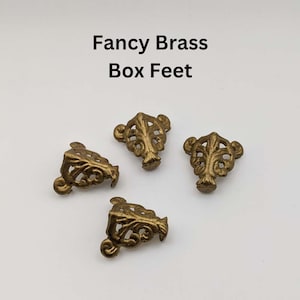 May include: Four ornate brass box feet with a decorative leaf design. The feet are a warm gold color and are arranged on a white surface. The text "Fancy Brass Box Feet" is above the feet.