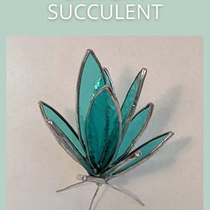 May include: A stained glass succulent sculpture featuring teal-colored glass petals framed in silver solder. The piece is labeled UPRIGHT SUCCULENT and SAGE GLASSWORK. The structure stands on thin, wire-like silver metal legs extending from the base.