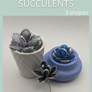 May include: Three stained glass succulent sculptures in varying shades of purple, blue, and dark teal, featuring metallic soldered edges. One is placed in a white textured pot, another in a light blue bowl, and one stands on a thin metal stem.