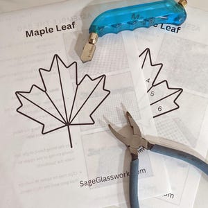 May include: A craft setup featuring a blue glass cutter, pliers, and printed paper patterns. The patterns include a maple leaf design with the text "Maple Leaf" and "SageGlasswork". The tools are for glass art projects.