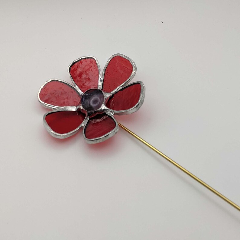 May include: A handcrafted red and purple flower pin with a gold-colored stem. The flower petals are a vibrant red, with a darker purple center. The pin is made of glass and metal, and is set against a white background.