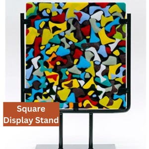 May include: A square glass art piece with a colorful abstract design in shades of red, yellow, blue, green, white, and black. The piece is mounted on a black metal stand.