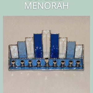 May include: A stained glass menorah with a beveled design. The menorah features blue, white, and clear glass panels, with silver candle holders. The text "BEVELED MENORAH" is at the top, and "SAGE GLASSWORK" is at the bottom.
