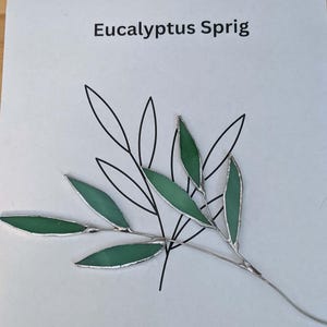 以下が含まれることがあります： 緑色のガラスの葉と銀色の金属アクセントが特徴の手作りのユーカリの小枝。 ユーカリの葉の黒い線画が描かれた白い背景に配置されています。 テキスト「Eucalyptus Sprig」が表示されています。