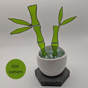 May include: A stained glass bamboo plant with bright green stalks and leaves, in a white ceramic pot filled with clear glass stones. The pot sits on a dark grey, hexagonal base. A green circle with "PDF pattern" is in the lower left.
