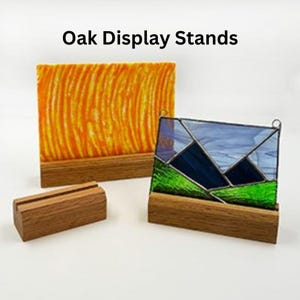 May include: Two oak display stands, one holding a stained glass piece with a blue and green mountain scene, and the other holding a piece of orange glass with a wavy pattern. The stands are made of natural wood and have a simple, elegant design. Oak Display Stands.