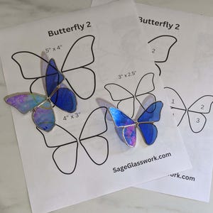 May include: Two stained glass butterfly ornaments with iridescent and blue wings rest on papers labeled "Butterfly 2". The larger butterfly measures 4" x 3", the smaller one is 3" x 2.5". The website SageGlasswork.com is visible.