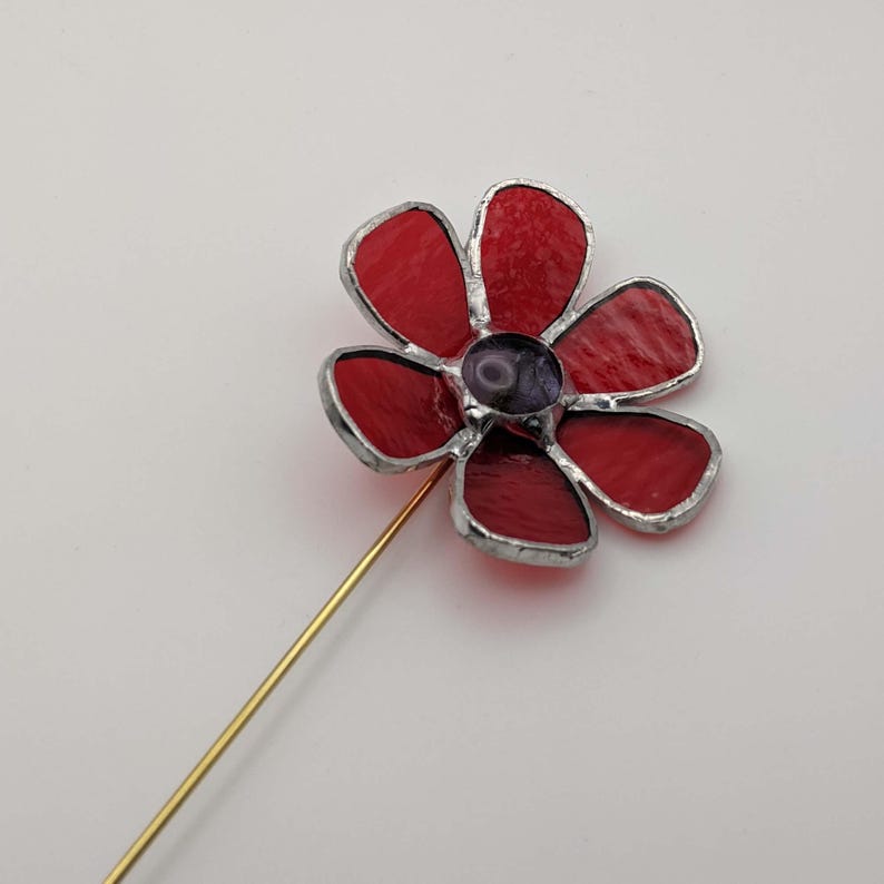 May include: A handcrafted red stained glass flower pin with a gold-colored stem. The flower has five petals with a dark purple center. The edges of the petals are outlined in silver.