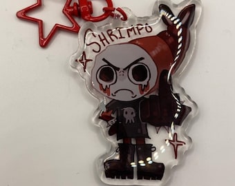 May include: A clear acrylic keychain featuring a cartoon character with a red shrimp-like headpiece and the word "SHRIMPO". The character has a skull on its chest and is wearing black boots. A red star-shaped charm is attached.