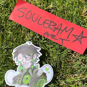 May include: A sticker featuring a stylized anime-style character with green and black hair, glasses, and a green outfit. The sticker has the word "MAC" at the bottom. A red rectangle with the words "SOULERAM ART" is also in the image.
