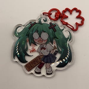 Pode incluir: Um chaveiro de acr&iacute;lico transparente com um personagem de desenho animado com cabelo verde, &oacute;culos e uniforme escolar, segurando uma placa que diz "SINNER". O chaveiro tem um fecho vermelho em forma de flor.