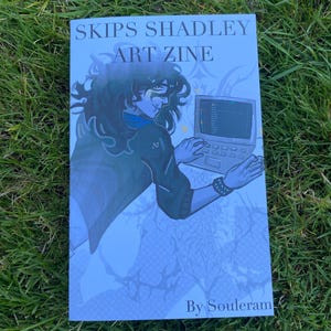 May include: A zine titled "SKIPS SHADLEY ART ZINE" featuring an illustration of a person working on a computer. The cover art is in shades of blue and white, with the text "By Souleram" at the bottom. The zine is lying on a bed of green grass.