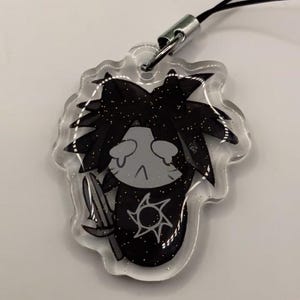 May include: A clear acrylic charm featuring a black and gray cartoon character with a spiky hairstyle, holding a trident. The charm has a silver-colored metal clasp and a black lanyard. The character has a sun symbol on its chest.