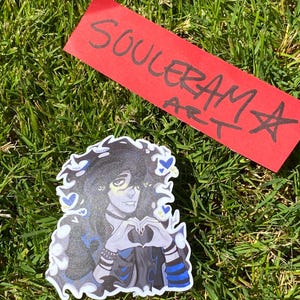 May include: A sticker featuring a stylized illustration of a person with dark hair and blue accents, forming a heart shape with their hands. The sticker is on a bed of green grass, with a red tag that reads "SOULERAM ART".