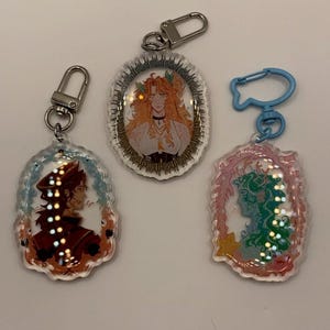 May include: Three clear acrylic keychains with character illustrations. The keychains feature oval frames with detailed portraits in shades of red, orange, and green. Each keychain has a different colored clasp.