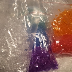 May include: Four bags of clear plastic beads, each containing a different colour: clear, purple, orange, and red.