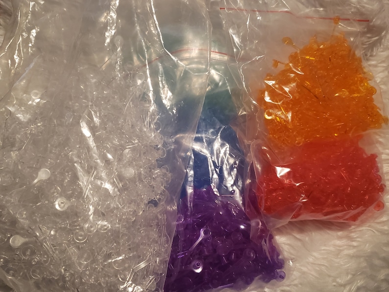 May include: Four bags of clear plastic beads, each containing a different colour: clear, purple, orange, and red.