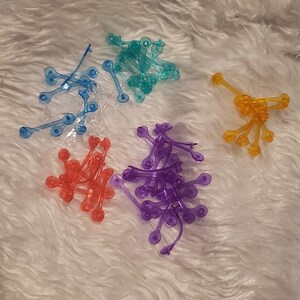 May include: Assortment of colourful plastic hair ties in blue, purple, orange, and clear. The hair ties are in a pile on a white fluffy surface.