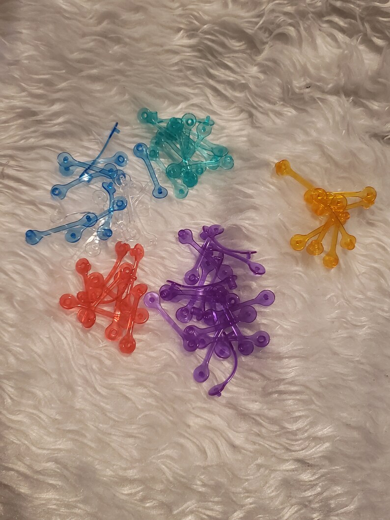May include: Assortment of colourful plastic hair ties in blue, purple, orange, and clear. The hair ties are in a pile on a white fluffy surface.