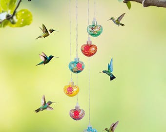 May include: Solar wind chime with hummingbird feeders. The wind chime features six colorful, glass-like orbs with floral designs, suspended from a brown circular top. Several hummingbirds are shown flying around the wind chime against a blurred green background.