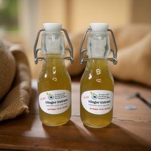 May include: Two small glass bottles filled with a golden liquid, labeled "Pure Ginger Extract." Each bottle has a white stopper and a metal clasp. The labels include the text "JC Farms of NC" and "Net Weight: 4 fl oz/118.29g."