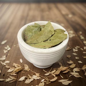 May include: A white ceramic bowl filled with dried bay leaves. The bowl is sitting on a wooden surface with scattered dried leaves.
