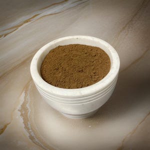 May include: A white ceramic bowl filled with a brown powder. The bowl has a textured surface and is sitting on a light brown surface.