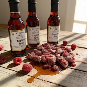 May include: Three bottles of raspberry syrup with black caps and labels that read "JC Farms of NC Raspberry Syrup." Fresh raspberries and a pool of syrup are on a wooden surface.