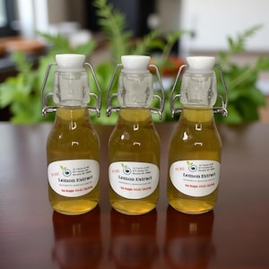 May include: Three small glass bottles filled with yellow liquid, each sealed with a white stopper and metal clasp. The bottles have labels that read "Lemon Extract." The background features green foliage and a dark brown surface.