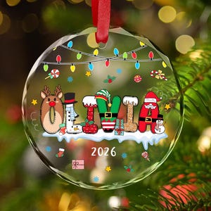 May include: A round, clear glass Christmas ornament featuring the name OLIVIA in colorful, holiday-themed letters. The design includes a string of multi-colored lights, small gift boxes, candy canes, and the year 2026 printed in white at the bottom.