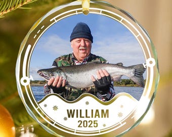 May include: A clear, round Christmas ornament with a photo of a person holding a fish. The ornament has a gold border and the name "WILLIAM" and the year "2025" printed on it. The ornament is hanging on a Christmas tree.
