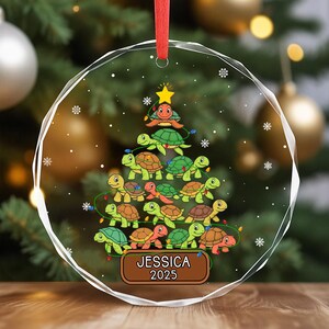 May include: A clear, round Christmas ornament with a red ribbon. The ornament features a tree made of cartoon turtles, with a star on top. The name "JESSICA" and the year "2025" are printed on a brown plaque at the bottom.