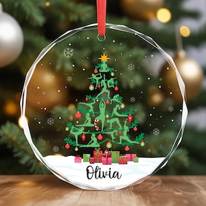 May include: A clear, round Christmas ornament with a red ribbon. The ornament features a green yoga tree design with red and green accents, snowflakes, and the name "Olivia" in black script. Small, colorful gift boxes are at the base.