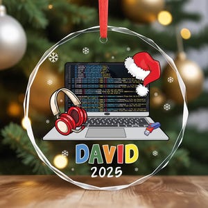 May include: Clear, faceted Christmas ornament with a red ribbon. The ornament features a laptop displaying code, a Santa hat, and red headphones. The name "DAVID" is written in colorful letters, with the year "2025" below.