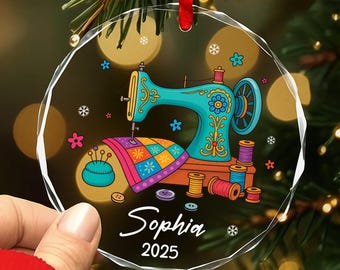 May include: A clear, round Christmas ornament featuring a teal sewing machine with floral details, a colorful quilt, and spools of thread. The ornament has the name "Sophia" and the year "2025" printed on it. A red ribbon is attached for hanging.