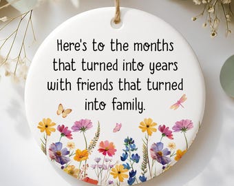 May include: A white, round ceramic ornament with a floral design and a jute string for hanging. The ornament features the text "Here's to the months that turned into years with friends that turned into family."
