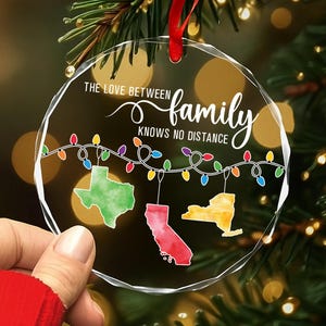 May include: A clear, round Christmas ornament with the text "THE LOVE BETWEEN family KNOWS NO DISTANCE." The ornament features colorful state outlines of Texas, California, and New York, connected by a string of lights.