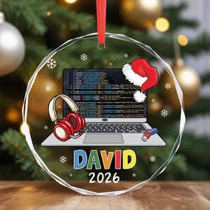 May include: A circular glass ornament featuring a laptop displaying code, red headphones, a USB drive, and a Santa hat. The name DAVID is printed in multicolored letters above the year 2026. A red ribbon is attached to the top for hanging.