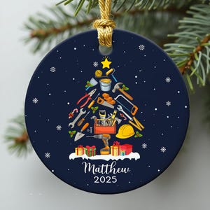 May include: A navy blue Christmas ornament with a gold string. The ornament features a Christmas tree design made of tools. It has the name "Matthew" and the year "2025" printed on it, with snowflakes and a star.