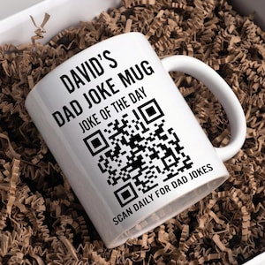 May include: A white ceramic mug featuring black text that reads DAVID'S DAD JOKE MUG, JOKE OF THE DAY, and SCAN DAILY FOR DAD JOKES, along with a large black and white QR code printed on the front surface of the mug for scanning purposes.