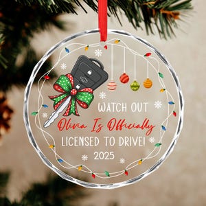 May include: Clear, round Christmas ornament with a car key design and a red ribbon. The key has a green and red bow. Text reads "Watch Out Olivia Is Officially Licensed to Drive! 2025". Decorative lights and ornaments surround the text.